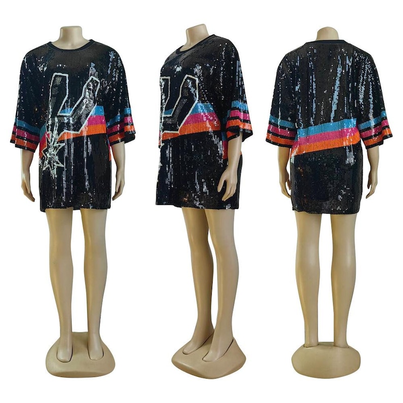 May include: Black sequined tunic dress with short sleeves. The dress features a white graphic design with a star and colorful stripes in pink, orange, and blue. The sleeves have matching stripes. The dress is displayed on a mannequin.