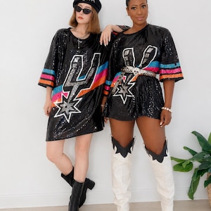 May include: Two people wearing black sequined dresses with a lightning bolt and star design. One person wears a black beret with "CHA" on it. The other person wears a belt with a skull buckle and white cowboy boots.