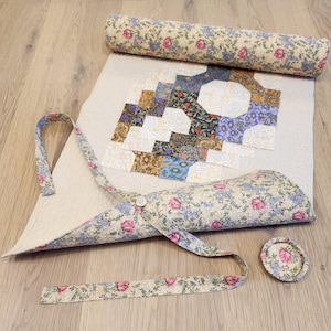May include: A beige fabric quilting project with a floral pattern. The project is rolled up and tied with a floral ribbon. The fabric is cut into squares and sewn together to create a patchwork design.