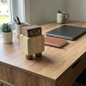 Könnte beinhalten: Eine Holzroboter-Skulptur mit rechteckigem Kopf und Körper sowie gelben Kugelfüßen steht auf einem Holzschreibtisch. Ein Laptop, ein Notizbuch und eine Tasse mit Stiften befinden sich ebenfalls auf dem Schreibtisch. Eine kleine Sukkulente im Topf ist sichtbar.