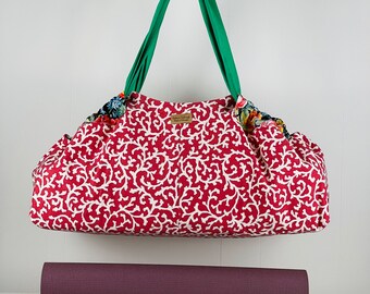 May include: A red and white patterned yoga bag with green straps. The bag is made of fabric and has a small label that says "Happy Yoga". A burgundy yoga mat is in the foreground.