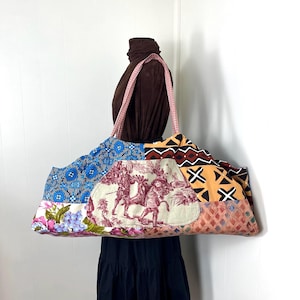 May include: A large patchwork tote bag with a variety of fabric patterns, including floral, geometric, and toile scenes. The bag has pink gingham straps and a brown interior. The bag is held by a mannequin.
