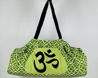 Puede incluir: Bolsa de yoga verde y blanca con un símbolo Om negro en la parte delantera. La bolsa tiene un cordón de ajuste negro y un asa negra.