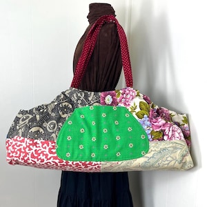 May include: A large patchwork tote bag with a variety of fabric patterns, including floral, geometric, and paisley designs. The bag features a green pocket with white flower accents and a red polka-dot strap. The bag is approximately 20 inches wide.