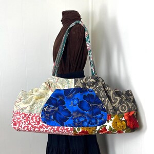 May include: A large, patchwork tote bag with a variety of fabric patterns and colors. The bag features a central blue panel with a floral design, surrounded by cream, red, and black patterned fabrics. The handles are green and floral.