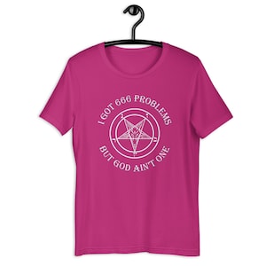 May include: A magenta t-shirt with a white graphic featuring a pentagram and the text "I GOT 666 PROBLEMS BUT GOD AIN'T ONE." The shirt is hanging on a black hanger.