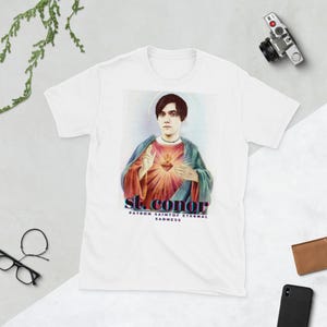 Può includere: Maglietta bianca con un'immagine colorata di una persona con un cuore e il testo "st. conor patron saint of eternal sadness".