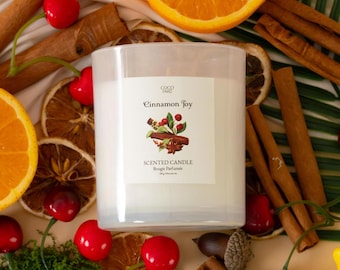 Può includere: Un barattolo di candela bianco con un'etichetta che recita "Coco Taro Cinnamon Joy Scented Candle Bougie Parfumée 200g/7oz net wt." La candela è circondata da bastoncini di cannella, fette di arancia essiccate e ciliegie rosse.