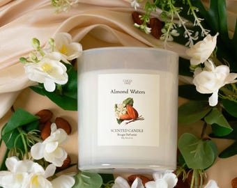 Può includere: Una grande candela profumata bianca, etichettata "Almond Waters", con un'illustrazione floreale e di mandorle. La candela è circondata da fiori bianchi, foglie verdi e mandorle sparse. Lo sfondo è un tessuto morbido color crema.