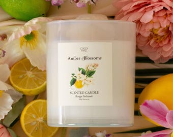 Può includere: Un barattolo di candela bianco con un'etichetta bianca che dice "Coco Tart Amber Blossoms Scented Candle Bougie Parfumee 200g 7oz net wt." La candela è circondata da limoni, lime e fiori rosa e bianchi.