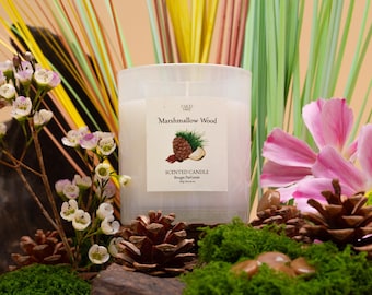 Può includere: Una candela bianca con un'etichetta che recita "Marshmallow Wood Scented Candle Bougie Parfumée". La candela è circondata da pigne, muschio e fiori.