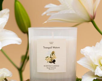 Può includere: Candela profumata bianca in un barattolo di vetro con la scritta "Tranquil Waters" e un'illustrazione floreale. Circondata da gigli bianchi e verde, posta su una superficie di legno.