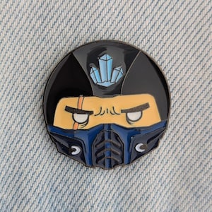 May include: A round enamel pin featuring a stylized character face with a black helmet, blue mask, and yellow skin. The pin has a blue crystal design on top and is set against a denim background.