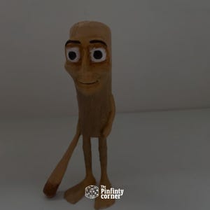 May include: A tan-colored, cartoonish figurine with a smiling face, large eyes, and a slender body. The figure has long, thin legs and arms, with one arm extended. The base of the figurine has the text "The Pinfinty corner".