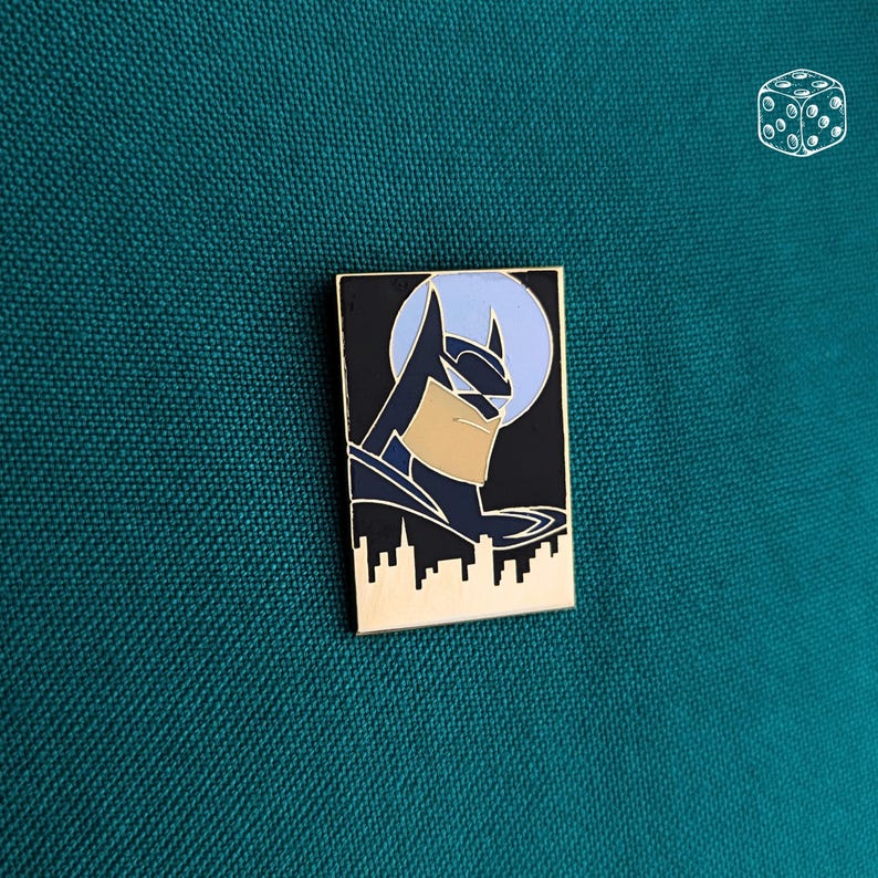 Pu&ograve; includere: Spilla rettangolare di Batman in smalto con bordo dorato. La spilla presenta il profilo di Batman su sfondo nero con una luna azzurra. Il design include una silhouette del paesaggio urbano nella parte inferiore.
