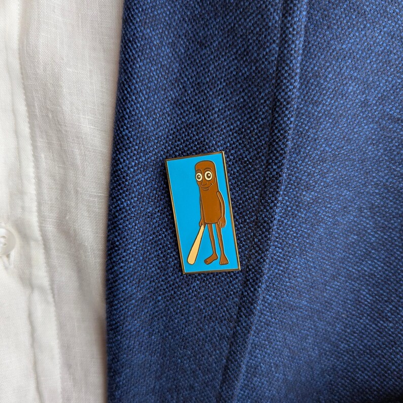 May include: A rectangular enamel pin featuring a cartoon character with a brown body, holding a baseball bat, set against a light blue background. The pin is attached to a navy blue blazer, with a white shirt visible in the background.