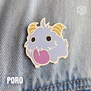 May include: A light blue enamel pin featuring a cartoon Poro character with a pink tongue and yellow horns. The pin has a white outline and the word "PORO" is printed in white at the bottom. The pin is set against a denim background.