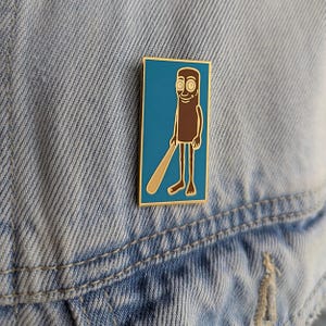 May include: A rectangular enamel pin featuring a cartoon character holding a baseball bat. The character is brown with a tan bat, set against a teal background with a gold border. The pin is attached to a denim jacket.