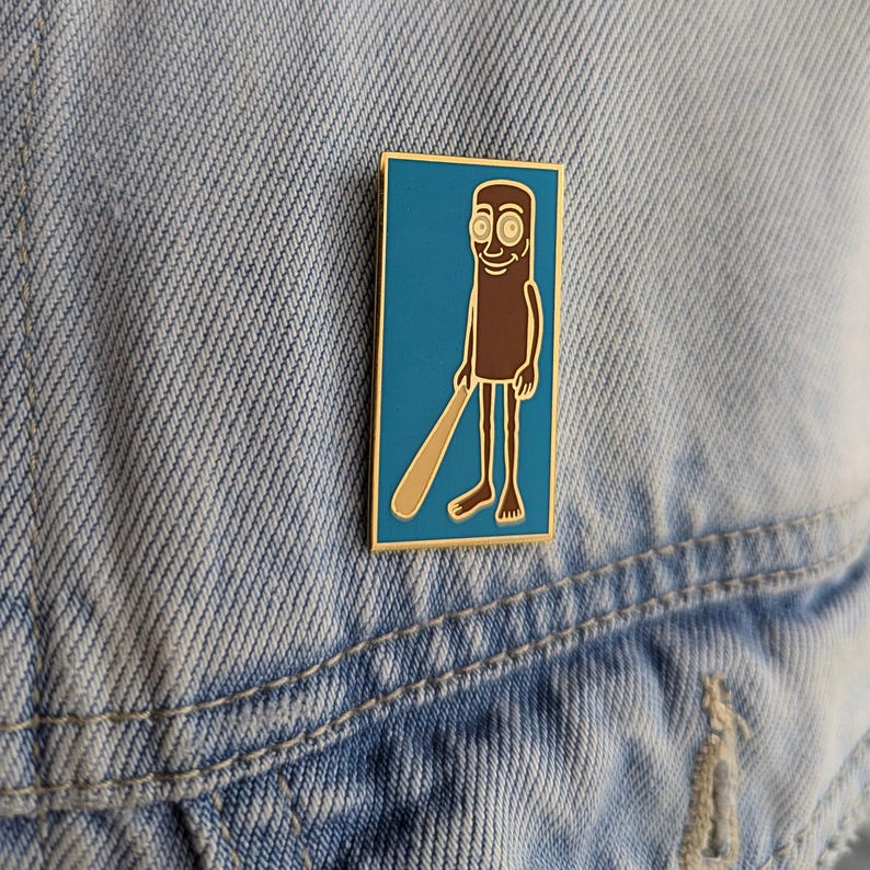 May include: A rectangular enamel pin featuring a cartoon character holding a baseball bat. The character is brown with a tan bat, set against a teal background with a gold border. The pin is attached to a denim jacket.