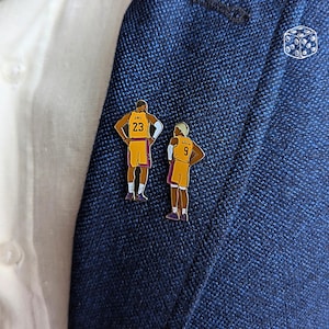 Könnte beinhalten: Zwei Emaille-Anstecknadeln mit Basketballspielern in gold-violetten Uniformen. Eine Nadel zeigt einen Spieler mit der Nummer 23, die andere einen Spieler mit der Nummer 9. Die Nadeln sind an einem marineblauen Blazer befestigt.