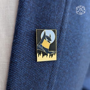 Pu&ograve; includere: Spilla rettangolare in smalto di Batman con bordo dorato. La spilla presenta un'immagine stilizzata della testa e della silhouette di Batman contro una luna azzurra e un paesaggio urbano nero. La spilla &egrave; attaccata a un blazer blu.