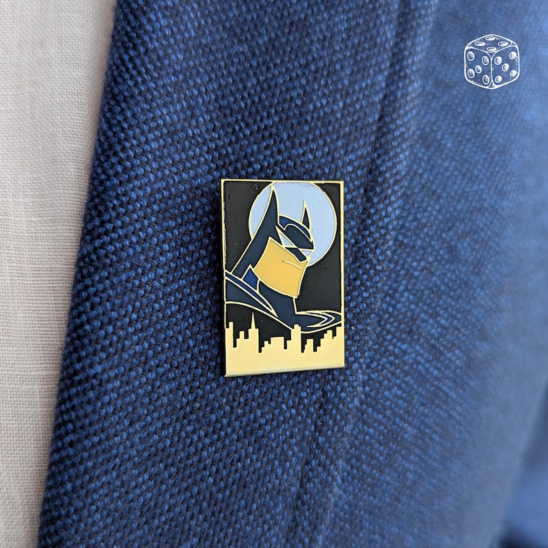 Pu&ograve; includere: Spilla rettangolare in smalto di Batman con bordo dorato. La spilla presenta un'immagine stilizzata della testa e della silhouette di Batman contro una luna azzurra e un paesaggio urbano nero. La spilla &egrave; attaccata a un blazer blu.