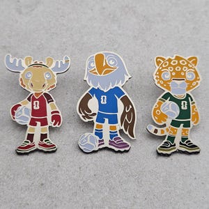 May include: Three enamel pins featuring anthropomorphic animal athletes: a moose in a maroon jersey, an eagle in a blue jersey, and a leopard in a green jersey. Each character holds a soccer ball and wears matching shorts and athletic shoes.
