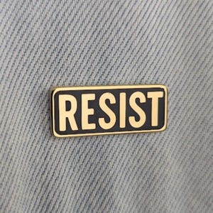 Op de afbeelding: Een rechthoekige speld met het woord "RESIST" in gouden letters op een zwarte achtergrond. De speld heeft een gouden rand en is geplaatst op een lichtblauwe denim stof.
