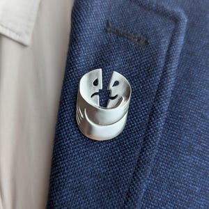 May include: A silver pin with two faces, one smiling and one frowning, intertwined in a hug.