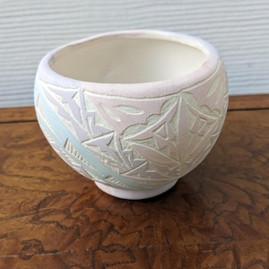 May include: A small, round ceramic bowl with a matte finish in soft pastel shades of pink, light blue, and cream. The exterior features a carved, geometric relief pattern covering the surface, while the interior is a smooth, solid cream color.
