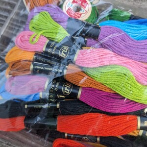 May include: A collection of Iris brand embroidery floss skeins in various colors including orange, lime green, pink, purple, blue, and black, packaged in a clear plastic bag. Each skein features a black paper label with the text 8 mts and Iris.