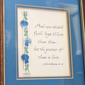 May include: Framed artwork with a blue mat and a wooden frame. The artwork features a quote from 1 Corinthians 13:13, with the words "And now abideth faith, hope, & love; these three: but the greatest of these is love." A decorative ribbon with blue flowers accents the left side.