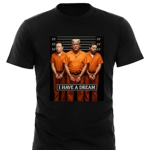 May include: Black t-shirt with a graphic of three men in orange jumpsuits and handcuffs. The men are standing in a line, and the text "I HAVE A DREAM" is printed below them.