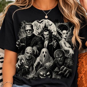 May include: A black t-shirt featuring a detailed monochromatic graphic print of classic horror movie monsters including Frankenstein's monster, Dracula, the Bride of Frankenstein, a mummy, a ghost, and a werewolf, with a castle and moon.