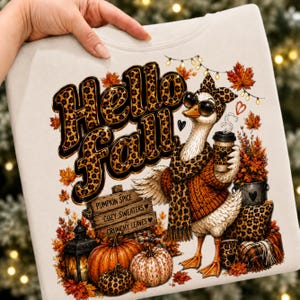 Könnte beinhalten: Ein cremefarbenes Sweatshirt mit einer Grafik einer Gans, die einen Schal und eine Sonnenbrille mit Leopardenmuster trägt und einen Kaffeebecher hält. Der Text lautet Hello Fall, Pumpkin Spice, Cozy Sweaters und Crunchy Leaves, umgeben von Herbstlaub.