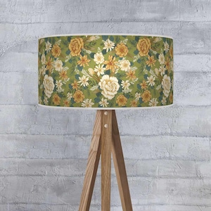 May include: A lampshade featuring a floral design with a green backdrop and yellow, white, and cream flowers. The lampshade is cylindrical and is supported by a wooden tripod lamp base.