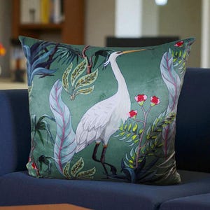 May include: Square cushion with a green background featuring a white heron and tropical foliage in shades of green, blue, and red. The cushion is placed on a blue sofa.