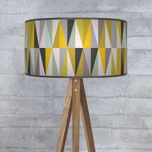 May include: A wooden tripod floor lamp with a geometric lampshade. The lampshade features a pattern of yellow, grey, black, and light blue triangles. The lamp has a natural wood finish and a grey trim.