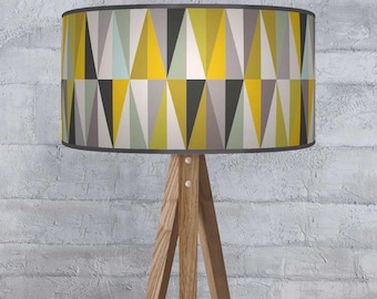 Può includere: Una lampada da terra a treppiede in legno con un paralume geometrico. Il paralume presenta un motivo di triangoli gialli, grigi, neri e azzurri. La lampada ha una finitura in legno naturale e un bordo grigio.