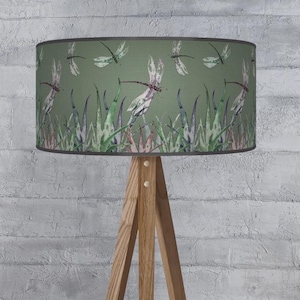 May include: A wooden tripod floor lamp with a green lampshade. The lampshade is decorated with a repeating pattern of dragonflies and tall grass. The dragonflies are white with black wings and red bodies. The grass is green, purple, and pink.