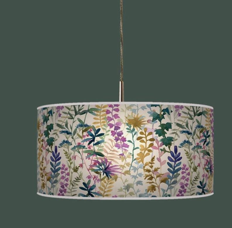 May include: A cylindrical pendant lamp with a floral design. The lampshade displays a watercolour pattern of diverse flowers and foliage in shades of purple, green, yellow, and blue against a cream backdrop. The lamp is suspended from a silver cord.