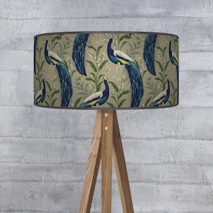 May include: A lampshade with a peacock design. The lampshade features a repeating pattern of blue, green, and yellow peacocks against a beige background with green foliage. The lampshade is mounted on a wooden tripod lamp base.