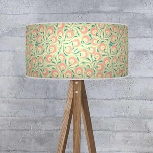 May include: A lampshade featuring a floral design in peach, teal, and cream hues. The cylindrical lampshade is mounted on a wooden tripod lamp base. The backdrop is a white brick wall.
