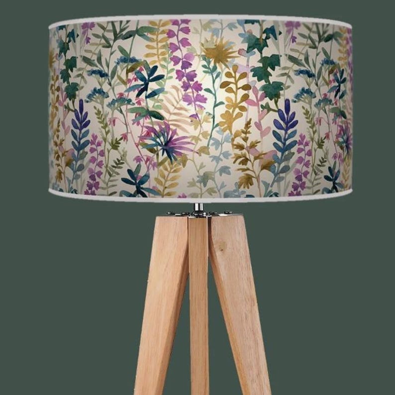 May include: A floor lamp with a light wood tripod base and a cylindrical lampshade. The lampshade displays a watercolour floral pattern in purple, blue, green, and yellow on a cream background. The lamp is set against a dark green backdrop.