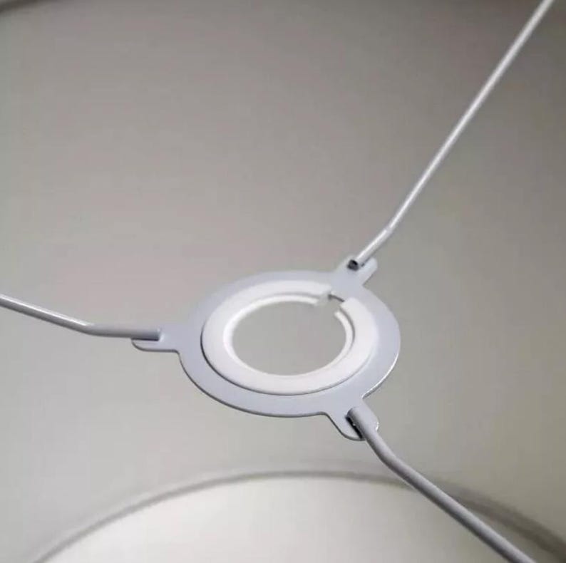 May include: A close-up shot of a grey lampshade frame. The frame has a circular design with a white inner ring and three wire supports. The metal frame is set against a soft, neutral background.