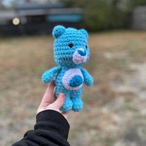 May include: A handmade, light blue crochet bear with a white belly and a blue heart-shaped design. The bear has small black eyes and a white muzzle. The arms and legs are also light blue. This plush toy is a cute, handcrafted item.