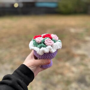 May include: A crocheted flower arrangement in a purple vase. The arrangement features red, pink, and green flowers with a white ruffled trim. The vase is held in a hand against a blurred outdoor background.