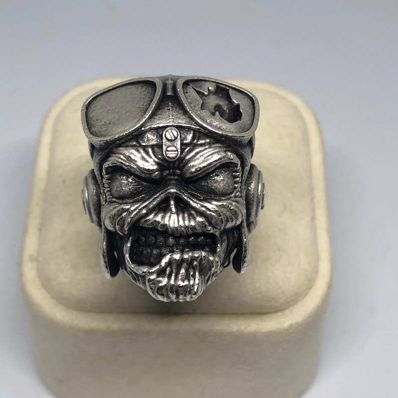 K&ouml;nnte beinhalten: Ein silberfarbener Ring mit einem detaillierten Totenkopf-Design, komplett mit Fliegerbrille und Kopfh&ouml;rern. Der Totenkopf hat einen bedrohlichen Ausdruck mit sichtbaren Z&auml;hnen. Der Ring ist wahrscheinlich aus Metall und wird auf einer neutralen Oberfl&auml;che pr&auml;sentiert.