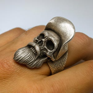 Può includere: Un anello a forma di teschio in argento con un design dettagliato. Il teschio ha una lunga barba e indossa un cappello. L'anello è su un dito, mostrando l'intricata lavorazione artigianale.