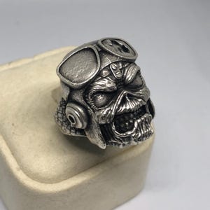 K&ouml;nnte beinhalten: Ein silberfarbener Ring mit einem detaillierten Totenkopf-Design, der eine Fliegerbrille tr&auml;gt. Der Sch&auml;del hat einen bedrohlichen Ausdruck mit sichtbaren Z&auml;hnen. Der Ring ist wahrscheinlich aus Metall und ein einzigartiges Schmuckst&uuml;ck.